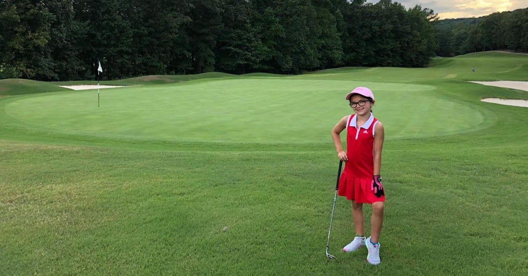 little girl in a red golf outfit standing by the green on a golf course with a putter