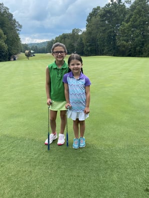 two little girls standing next to the green on a golf course with their putters